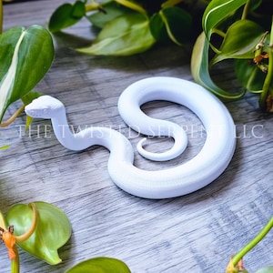 May include: A solid white snake figurine coiled in a circular shape with its head raised. The surface of the snake features a detailed scale texture. The text THE TWISTED SERPENT LLC is printed in white across the center of the image.