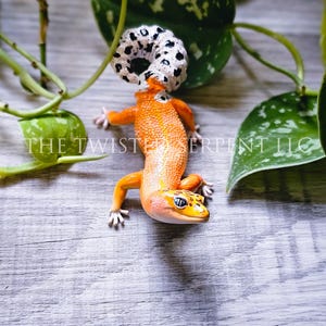May include: An orange and yellow gecko figurine with a white and black spotted ring around its head. The gecko is posed on a wooden surface, with green leaves and the text "THE TWISTED SERPENT LLC" in the background.
