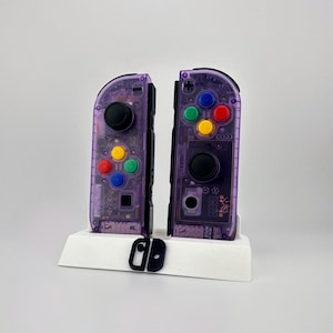 May include: Two purple Nintendo Switch Joy-Con controllers with visible internal components, featuring black joysticks and colorful buttons. They are displayed on a white stand, with a black rail attachment in front.