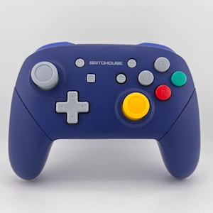 May include: A navy blue video game controller with a variety of colored buttons. The controller has a gray joystick, a directional pad, and several buttons in red, green, and yellow. The text "SWITCHOUSE" is printed on the top.