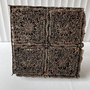 Op de afbeelding: Een vierkante houten batikstempel met een ingewikkeld bloemen- en bladerenpatroon. De stempel is donkerbruin met lichtbruine details en het ontwerp is symmetrisch. De randen van de stempel zijn licht versleten, wat tekenen van gebruik vertoont.