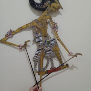 Op de afbeelding: Een traditionele Indonesische Wayang Kulit-pop. De pop is gemaakt van ingewikkeld gesneden en beschilderd leer, met een gedetailleerd ontwerp met gouden, rode, blauwe en witte accenten. De pop wordt vastgehouden door zwarte staven en de figuur heeft een kenmerkende hoofdtooi.