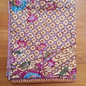Puede incluir: Una pieza de tela rectangular con un patrón estilo batik sobre un fondo de cuadrícula amarillo, motivos florales morados y detalles azules. El diseño incluye una sección inferior con patrones de remolinos marrones y flecos decorativos.