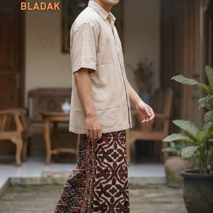 Könnte beinhalten: Ein Mann trägt ein beiges kurzärmeliges Hemd und einen gemusterten Sarong. Der Sarong hat ein braunes und cremefarbenes geometrisches Design. Das Wort "BLADAK" wird in orangefarbenem Text angezeigt. Das Bild enthält auch den Text "Saroeng Djawa".