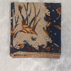 Puede incluir: Un cuadrado de tela doblado con un patrón batik, que presenta un centro color crema con líneas onduladas y motivos de hojas marrones, bordeado por una sección azul oscuro con elementos florales y un borde marrón a rayas en el dobladillo.
