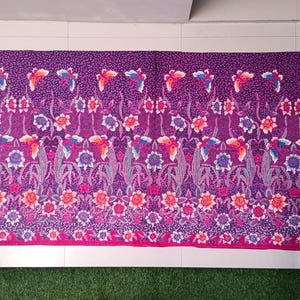 May include: A rectangular textile with a repeating floral and bird pattern in shades of purple, pink, and orange. The edges are trimmed with a solid magenta border. The design features stylized flowers and birds.
