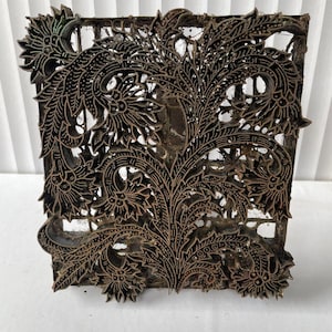 Op de afbeelding: Een vierkant, donkerbruin houten blok met een ingewikkeld bloemenontwerp. Het blok heeft een gedetailleerd patroon van bladeren, bloemen en wervelende lijnen, waardoor een decoratieve en sierlijke uitstraling ontstaat. Het ontwerp is in het hout gesneden, waardoor een reliëfeffect ontstaat.