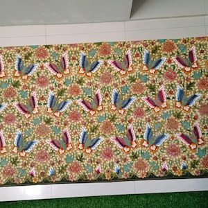 Op de afbeelding: Een rechthoekig textiel met een ingewikkeld bloemen- en vogelpatroon. Het ontwerp bevat herhaalde motieven van gestileerde vogels in blauw, rood en wit, tegen een achtergrond van bloemen in roze, oranje en groen. De randen zijn afgewerkt met een geschulpte rand.