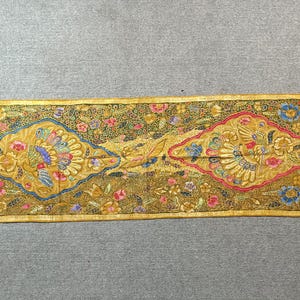 Könnte beinhalten: Ein rechteckiger, verzierter Stoff mit goldenem Rand und aufwendigen Mustern. Das Design zeigt zwei zentrale, rautenförmige Motive mit Vögeln und floralen Elementen in Gold, Blau, Rosa und Grün. Der Hintergrund ist dunkel olivgrün.
