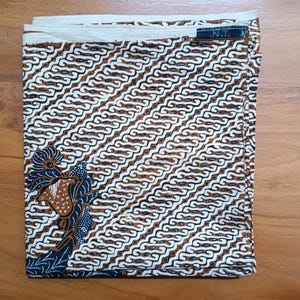 Könnte beinhalten: Ein gefalteter rechteckiger Stoff mit einem sich wiederholenden diagonalen Batikmuster in Braun, Weiß und Dunkelblau. Ein florales Motiv ist in der unteren linken Ecke sichtbar, und ein kleines dunkles Etikett mit dem Text N.T. befindet sich am oberen rechten Rand.