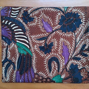 Op de afbeelding: Een vierkant stuk stof met een bruine achtergrond en een gedetailleerd batikpatroon. Het ontwerp bevat donkerblauwe bloemmotieven, paarse bladvormen en felgroene accenten, allemaal verbonden door ingewikkeld wit lijnwerk.