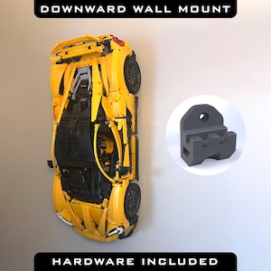 May include: A yellow Lego car model mounted on a wall with a black wall mount bracket. The text "DOWNWARD WALL MOUNT" is at the top of the image and "HARDWARE INCLUDED" is at the bottom of the image.