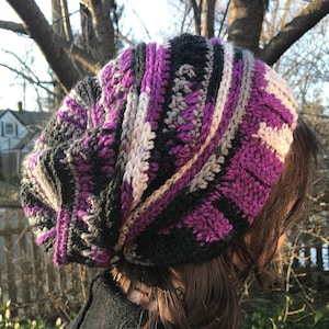 Pode incluir: Um chapéu de crochê feito à mão em estilo folgado. O chapéu apresenta um design de blocos de cores com listras pretas, roxas, cinzas e branco sujo. O chapéu é feito de fio macio e parece quente e confortável.