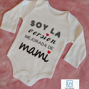 Puede incluir: Un body blanco de manga larga para bebé con texto negro que dice "Soy la versión mejorada de mami" con dos corazones rojos.