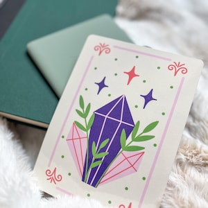 Könnte beinhalten: Eine cremefarbene Karte mit einem violetten Kristall, umgeben von grünen Blättern und rosa Diamantenformen. Die Karte hat einen rosa Rand mit grünen und roten Akzenten.