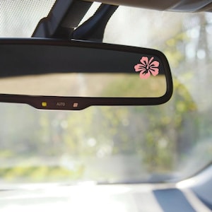 May include: A pink hibiscus flower decal on a rearview mirror in a car. The mirror has an "AUTO" button.