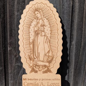 Pode incluir: Uma placa de madeira castanho-claro cortada a laser com uma grava&ccedil;&atilde;o de Nossa Senhora de Guadalupe. A base inclui o texto: Mi bautizo y primer a&ntilde;ito, Camila A. Lopez, 01.24.26. A placa tem uma borda recortada e fica em p&eacute;.