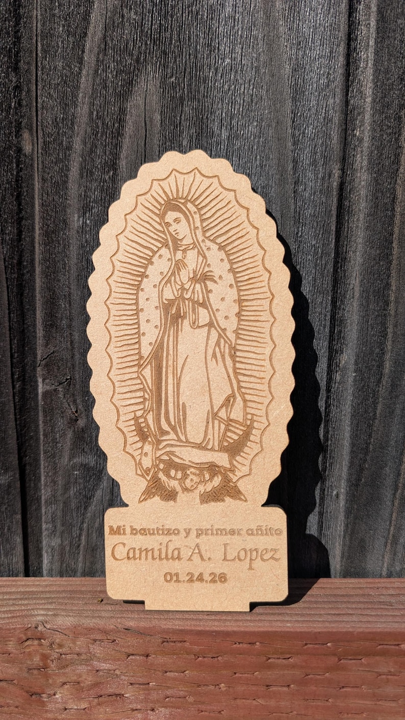 Pode incluir: Uma placa de madeira castanho-claro cortada a laser com uma grava&ccedil;&atilde;o de Nossa Senhora de Guadalupe. A base inclui o texto: Mi bautizo y primer a&ntilde;ito, Camila A. Lopez, 01.24.26. A placa tem uma borda recortada e fica em p&eacute;.