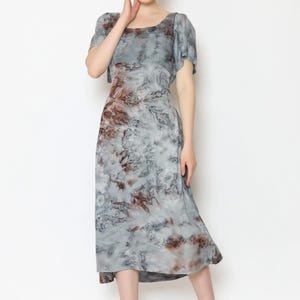 A person stands against a plain background wearing a stylish gray and brown tie back dress. The dress features short sleeves and a mid-length hem. They are smiling, touching their face with one hand, and wearing sandals.