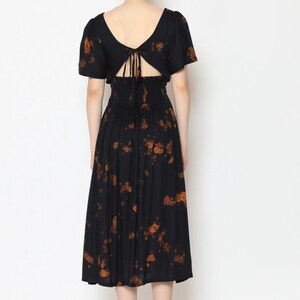 A woman is wearing a black tie back dress adorned with orange floral patterns. The dress features a low, open back with delicate tie details and puffed short sleeves. She stands on a plain white background, her hair in a bun, and complements the look with flat sandals.