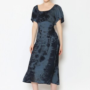 A person smiles, standing against a plain background in a striking tie-back dress. The blue-and-black tie-dye outfit features short sleeves and a square neckline. They complement the look with sandals, and their hair is neatly tied back.