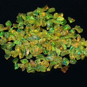 May include: A collection of raw, uncut opal gemstones. The opals are a mix of yellow, green, and orange hues, with a translucent quality. The gemstones are scattered on a black surface, creating a contrast that highlights their colors.