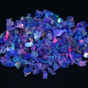 May include: A pile of rough-cut, iridescent blue opal gemstones. The stones have a fractured appearance with flashes of pink, green, and purple. The opals are scattered against a black background, highlighting their vibrant colors and textures.