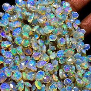 May include: A collection of faceted, pear-shaped opal gemstones featuring a translucent white base with vibrant flashes of blue, green, and orange fire. Each individual stone is polished to a high shine, reflecting light across the surface.