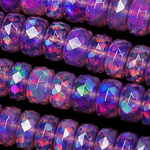May include: A collection of faceted, round beads arranged in horizontal rows. Each bead features a translucent, iridescent purple base with vibrant, multi-colored flashes of pink, blue, and green light reflecting across the geometric surfaces.