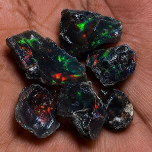 May include: A collection of five raw black opal stones with irregular shapes and dark, glossy surfaces. Each stone displays vibrant flashes of iridescent green and red colour play throughout the dark, textured mineral structure.