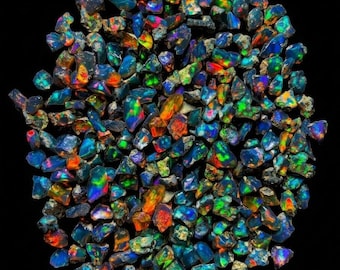 May include: A collection of small, raw black opal gemstones arranged in a circular cluster. Each stone features a dark base with vibrant, iridescent flashes of blue, green, orange, and red light reflecting from their irregular, multifaceted surfaces.