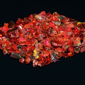 Puede incluir: Un montón de piedras preciosas de ópalo rojo y naranja en bruto. Los ópalos tienen un aspecto ardiente con destellos verdes y amarillos, sobre un fondo negro. Estas piedras preciosas son ideales para la fabricación de joyas.