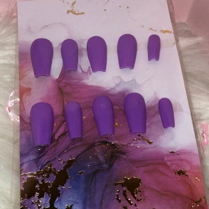 Puede incluir: Un juego de diez uñas postizas de color morado, mostradas en una tarjeta blanca con un diseño de mármol rosa y morado.