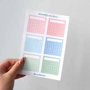 Op de afbeelding: Een vel met zes printbare maandelijkse trackers in pastelroze, blauw en groen. Elke tracker heeft een lege ruimte bovenaan voor een titel en een raster van vierkanten om data te markeren.