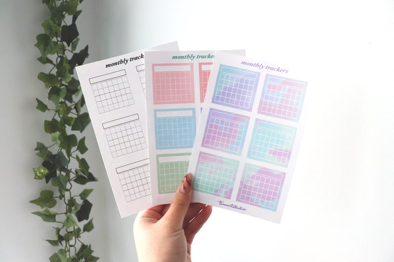 Op de afbeelding: Drie vellen met maandelijkse kalendertrackers. Het eerste vel is blanco, het tweede vel heeft pastelroze, blauwe en groene vierkanten, en het derde vel heeft pastelpaarse, blauwe en groene vierkanten met een aquareleffect.