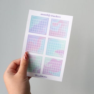 Op de afbeelding: Een vel met zes pastelkleurige maandelijkse kalendertrackerstickers. Elke sticker heeft een raster met een witte omtrek en een aquarelachtige achtergrond. De tekst "monthly trackers" staat bovenaan het vel gedrukt.