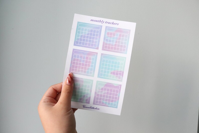 Op de afbeelding: Een vel met zes pastelkleurige maandelijkse kalendertrackerstickers. Elke sticker heeft een raster met een witte omtrek en een aquarelachtige achtergrond. De tekst "monthly trackers" staat bovenaan het vel gedrukt.