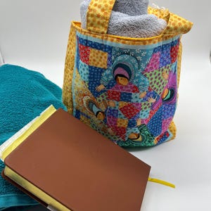 May include: A colorful tote bag with a patchwork design, holding rolled gray towels. A brown book with a yellow ribbon bookmark and a teal towel are also present. The bag has a yellow floral patterned handle and trim.