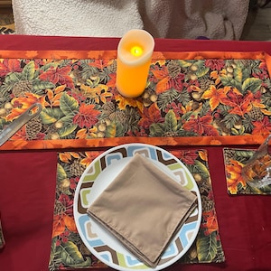 May include: A table setting with a fall theme. Includes a table runner, placemats, and coasters with a leaf and pinecone pattern. A lit candle, a plate with a napkin, and a glass are also present. The table is covered with a red tablecloth.