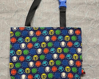 May include: A blue tote bag with a black strap and a blue buckle. The bag is patterned with colorful superhero logos in red, yellow, green, and blue. The bag is made of fabric.