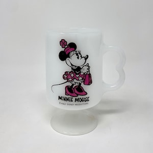 May include: White ceramic mug with a pink and black image of Minnie Mouse holding a shopping bag. The mug has a heart-shaped handle and a base. The text "Minnie Mouse" and "&copy; WALT DISNEY PRODUCTIONS" is printed on the mug.