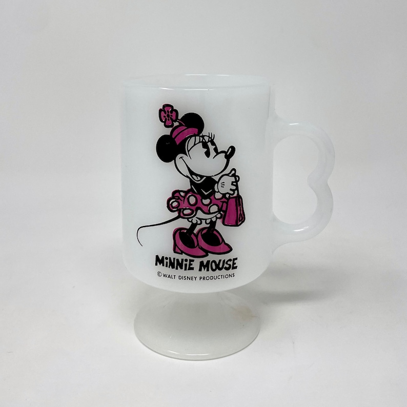 May include: White ceramic mug with a pink and black image of Minnie Mouse holding a shopping bag. The mug has a heart-shaped handle and a base. The text "Minnie Mouse" and "&copy; WALT DISNEY PRODUCTIONS" is printed on the mug.