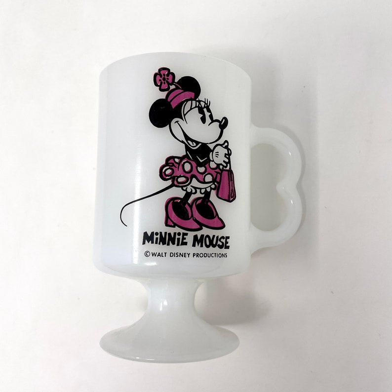 May include: White ceramic mug with a pink and black image of Minnie Mouse holding a purse. The mug has a heart-shaped handle and a pedestal base. The text "Minnie Mouse" and "&copy; WALT DISNEY PRODUCTIONS" is printed on the mug.