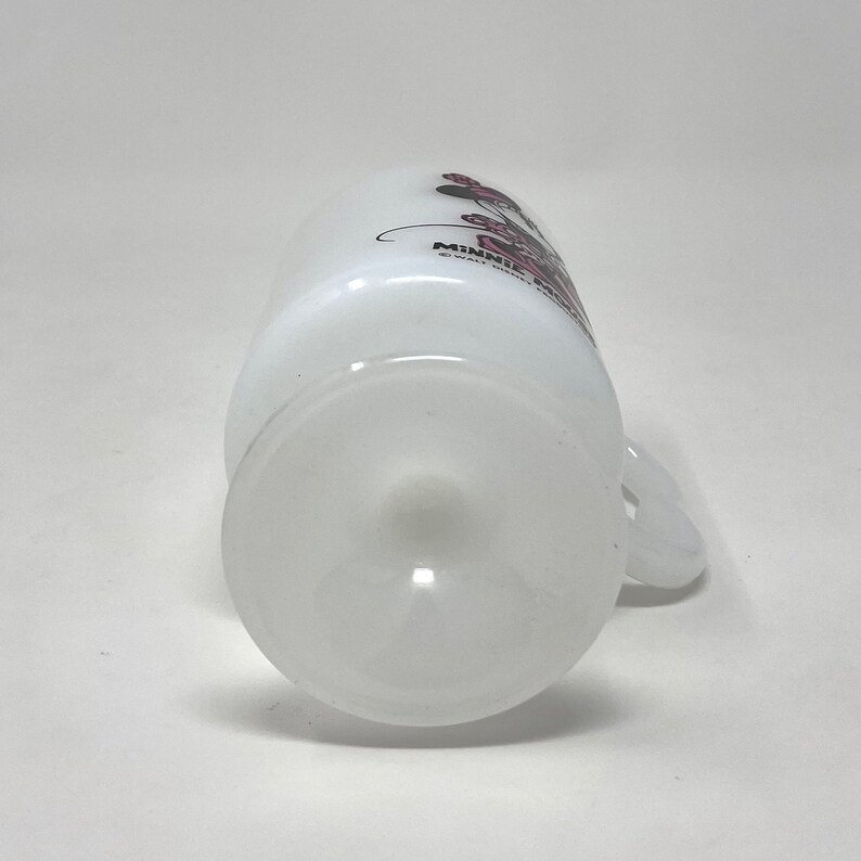 May include: White ceramic mug with a Minnie Mouse design. The mug has a handle and a white base.