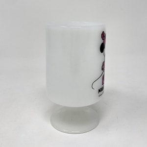May include: White ceramic mug with a Minnie Mouse illustration. Minnie is wearing a pink dress and a pink bow. The mug has a white pedestal base.