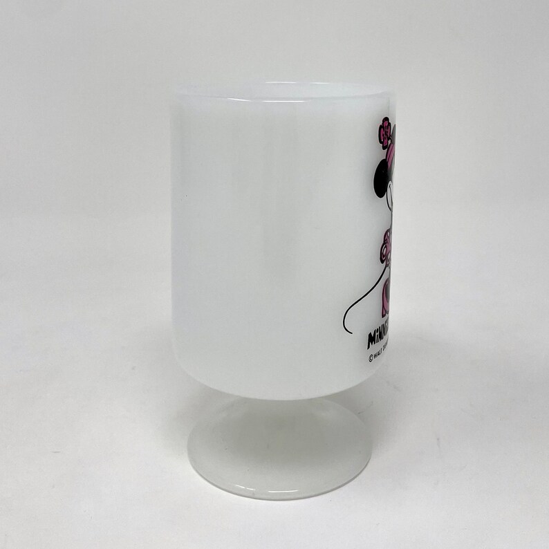 May include: White ceramic mug with a Minnie Mouse illustration. Minnie is wearing a pink dress and a pink bow. The mug has a white pedestal base.