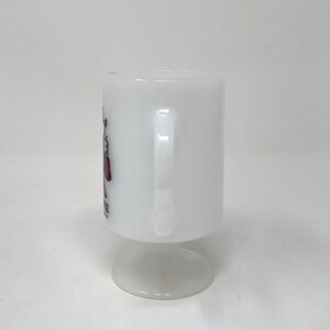 May include: White ceramic mug with a pink and black design. The mug has a handle and a pedestal base.