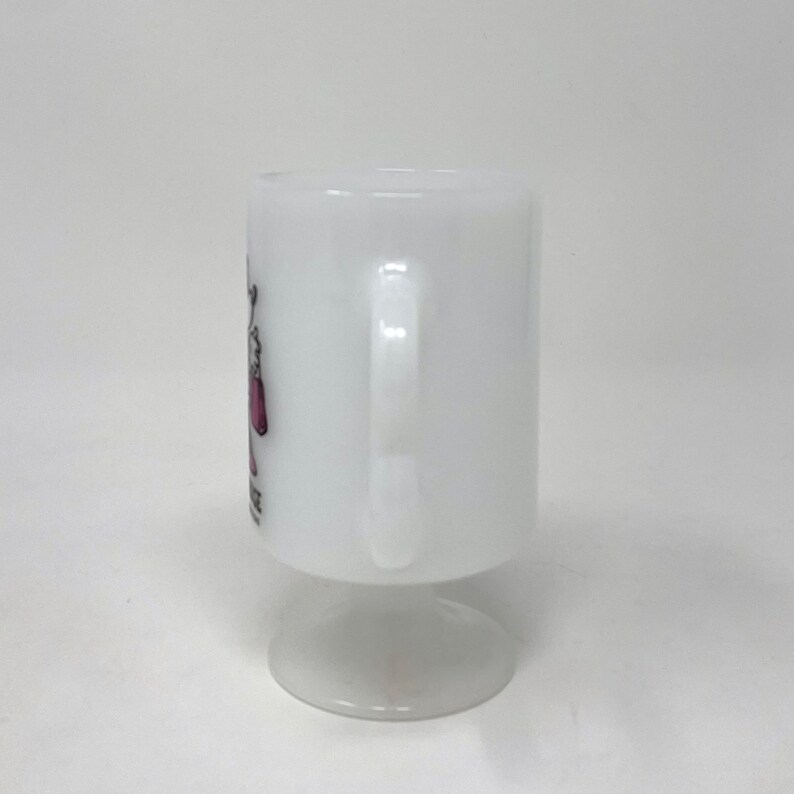 May include: White ceramic mug with a pink and black design. The mug has a handle and a pedestal base.