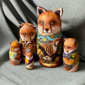 May include: A set of five hand-painted wooden nesting dolls, or matryoshka dolls, featuring foxes and woodland animals. The largest doll is a fox with a blue scarf and a brown and white bird. The other dolls are smaller foxes with different animal companions.