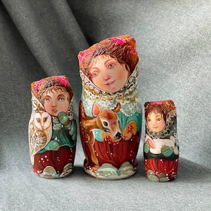 Pode incluir: Um conjunto de três bonecas russas de encaixe pintadas à mão, ou bonecas matryoshka. A boneca maior representa uma mulher com um vestido azul e verde segurando um veado. A boneca do meio representa uma mulher com um vestido verde e vermelho segurando uma coruja. A boneca menor representa uma mulher com um vestido vermelho e branco segurando um pássaro branco.
